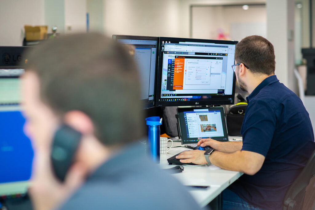 An IT technician working at a desk with multiple screens. Nearby, a blurred person is on the phone.