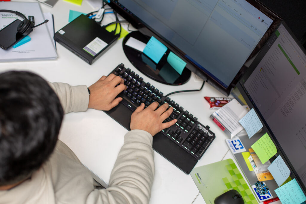 A person in a white sweater types on a keyboard at a desk with dual monitors. Sticky notes, a headset, and office supplies are scattered around.