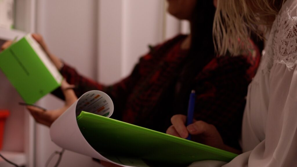 Two women are in a room holding a document and taking notes. One wears a red plaid shirt, the other a white lace blouse. The focus is on their hands.