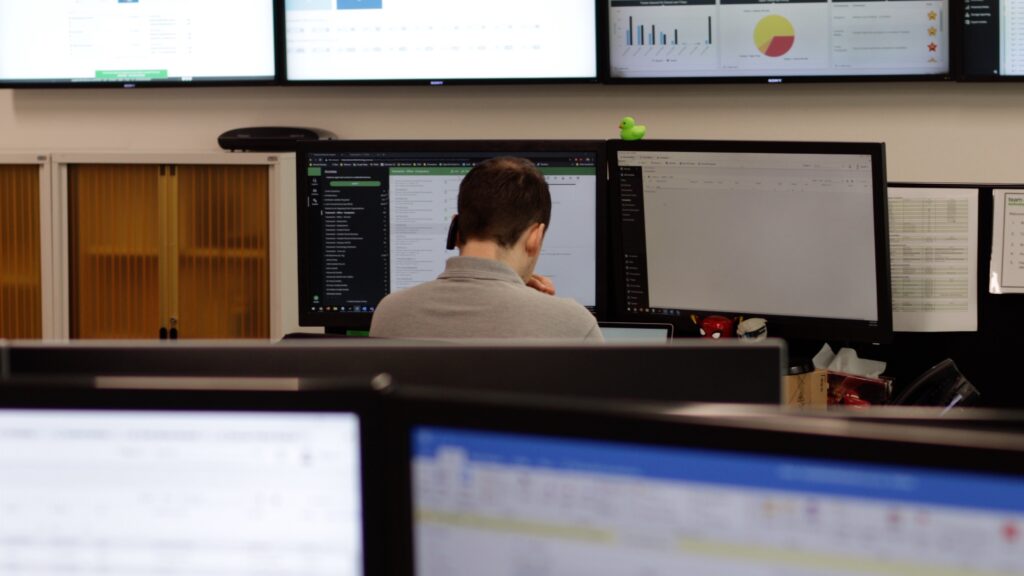 A person working at a desk with multiple computer monitors, displaying charts and spreadsheets in an office setting.
