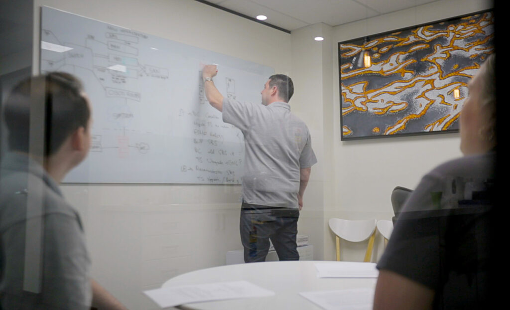 Teamwork Technology's founder James Borg writes on a whiteboard in a conference room, while two colleagues observe.