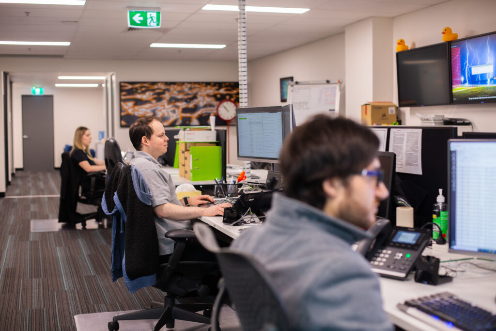 Teamwork Technology's open-plan office with three people working at computer desks. Monitors display data.